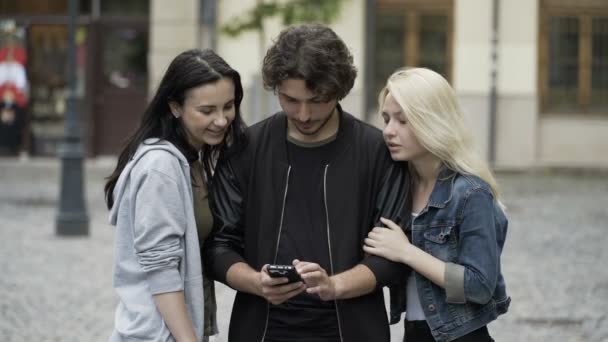 Dvě mladé hezké dívky dává překvapivě líbat muž tváře, zatímco on pomocí smartphone na městské ulici — Stock video