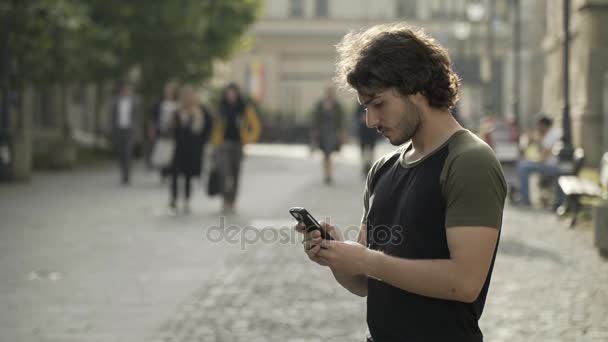 Bonito homem esperando em um lugar urbano público para amigos mensagens de texto no aplicativo de mídia social em seu smartphone — Vídeo de Stock