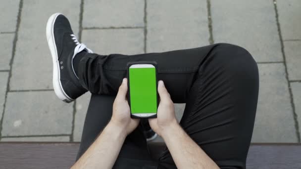 Casual geklede man zittend op de Bank in een park op zoek en het gebruik van zijn smartphone met Chromakey groen scherm — Stockvideo