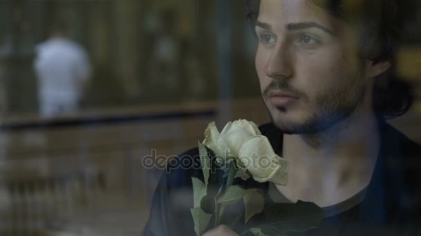 Nervózní mladý muž v lásce čekání na přítelkyni s květy sedí u okna v hospodě — Stock video