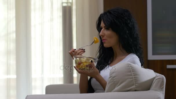 Молодая женщина сидит на диване, смотрит телевизор и ест фруктовый салат, питаясь здоровым завтраком — стоковое видео
