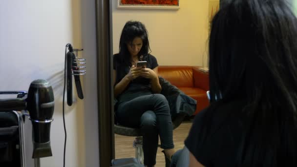 Chica joven sentada en la silla y mensajes de texto en el teléfono móvil en el salón de belleza — Vídeos de Stock