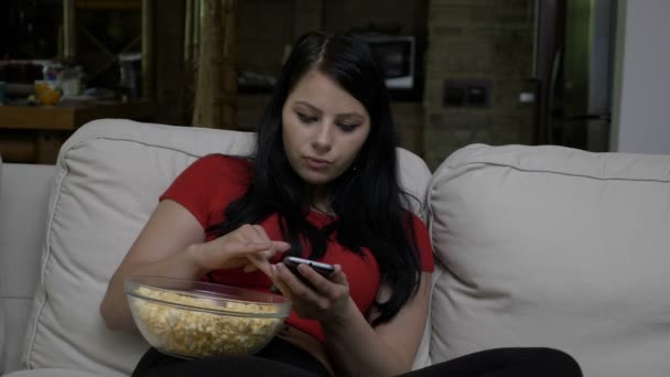 Kvinna sitter i soffan med en skål popcorn i hennes knä och textning på smart telefon under tv reklamavbrott — Stockvideo