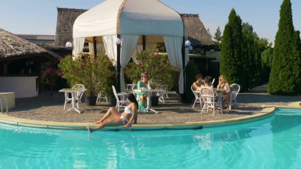 Persone che si rilassano a bordo piscina sedute a tavola su una terrazza e donne sedute sul bordo della piscina in una calda giornata estiva — Video Stock