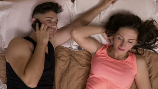 Joven hablando en el teléfono inteligente en la cama junto a su novia acariciando su cabello esperando a que pase tiempo juntos — Vídeos de Stock