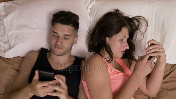 Pareja joven acostada en la cama charlando en las redes sociales adictos a los teléfonos inteligentes ignorándose mutuamente — Vídeos de Stock