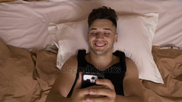 年轻的男人微笑着，躺在床上睡在社交媒体上使用智能手机前网络 — 图库视频影像