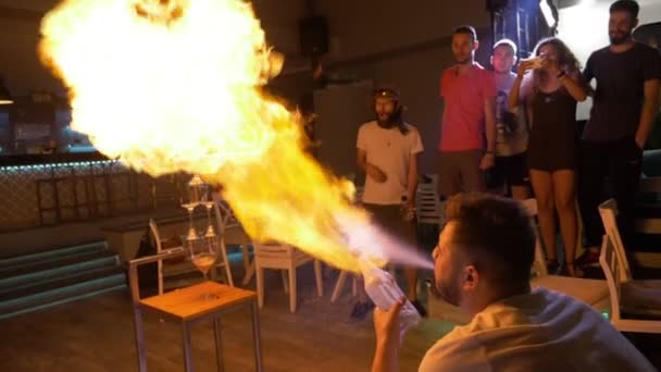 Homem fazendo truque pulverizando chama de vodka de conjuntos de garrafas em pilha de fogo de óculos — Vídeo de Stock