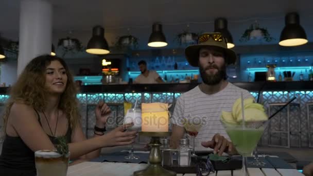Група молодих стильних друзів, які сидять за столом у клубному залі, насолоджуються алкогольними коктейлями, тостами та розважаються у вихідні — стокове відео