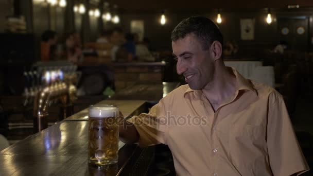 Мужчина сидит один в баре в переполненном пабе и пьет пиво — стоковое видео