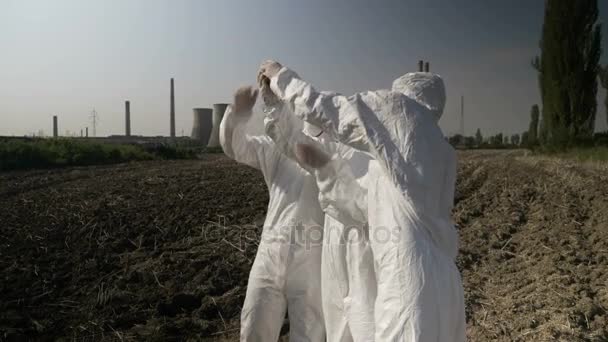 Biólogos trabajadores en ropa de materiales peligrosos mirando a un pedazo de tierra contaminada de un campo agrícola en la refinería — Vídeo de stock