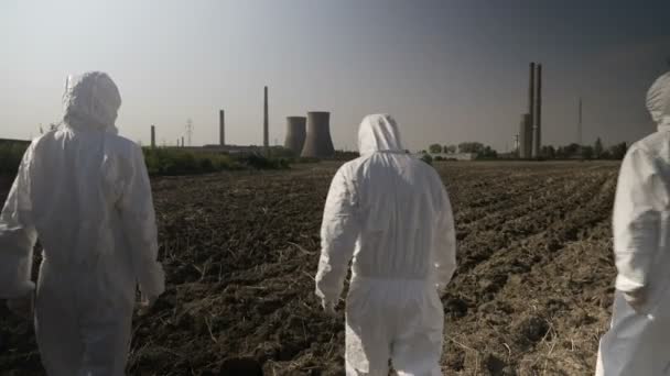 Equipe de ecologistas funcionários em ternos de perigo olhando para o ambiente contaminado e área agrícola caminhando em direção a uma refinaria de energia nuclear — Vídeo de Stock