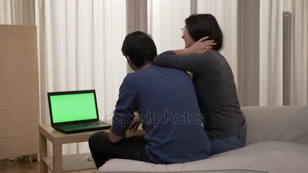 Усміхнена пара сидить на краю ліжка, дивлячись на ноутбук з зеленим екраном, маючи чудову розмову і показуючи прихильність — стокове відео