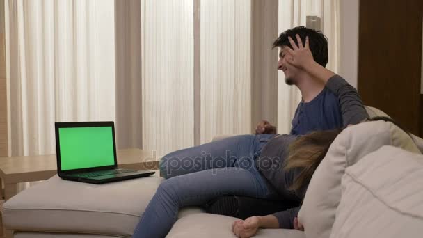 Забавная молодая пара обнимается и дурачится дома на белом диване во время просмотра фильма на ноутбуке с зеленым экраном целуется и проявляет привязанность — стоковое видео