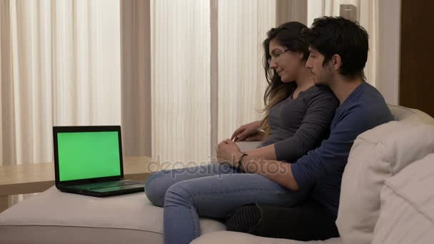 Молода жінка сидить на колінах свого хлопця на білому дивані вдома дивитися фільм на ноутбуці з зеленим екраном і обговорювати — стокове відео