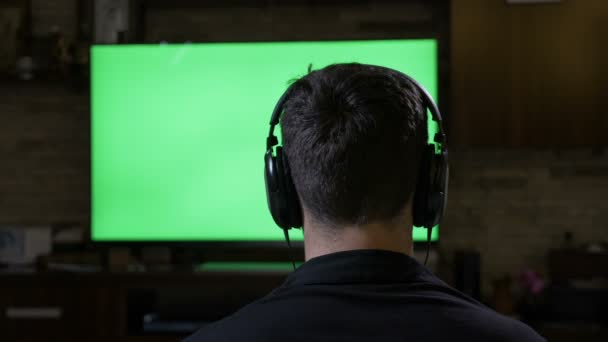 Relaksujący młody człowiek z słuchawki gry wideo przed chroma Klucz zielonego ekranu monitora tv — Wideo stockowe