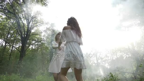 Two beautiful friends spin and dance in a meadow through smoke in slow motion — Stock Video