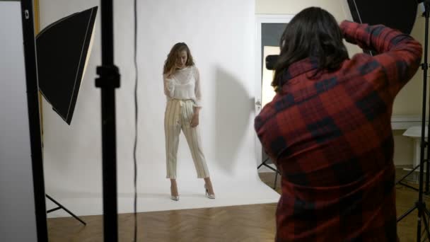 Schönes junges Model mit langen Haaren posiert in einem Studio mit weißem Hintergrund und überprüft dann ihre Fotos — Stockvideo