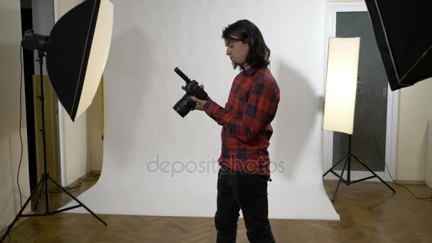Jonge aantrekkelijke witte mannelijke fotograaf zit in zijn de foto's van zijn digitale camera en tevreden van zijn werk controleren fotostudio — Stockvideo