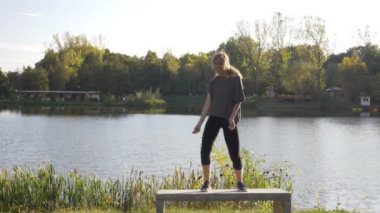 Dans ve doğa güneş ışığı yavaş çekimde bir göl yakınındaki parkta bir bankta kadar pozitif enerji yaymak mutlu fitness modeli kız