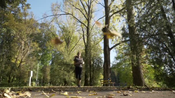 Skinny dospívající dívka jogging v parku, zatímco listí na podzim ze stromů na slunečný podzimní den v víkend v pomalém pohybu — Stock video