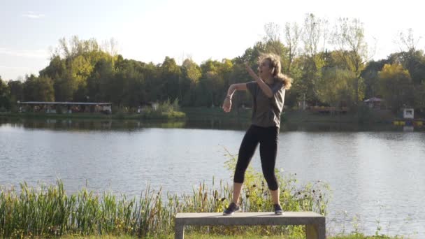 Приваблива щаслива молода студентка, насолоджується вихідним днем у парку, танцює біля озера і перевертає волосся повільним рухом — стокове відео