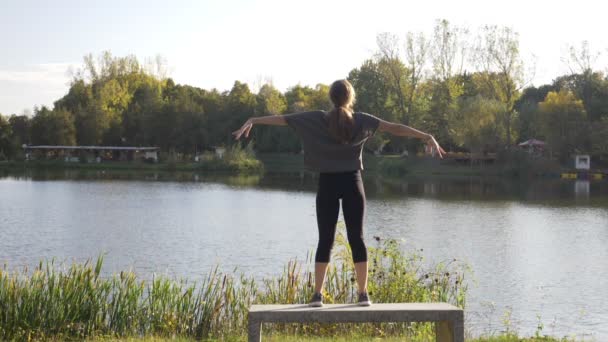 Menina atraente magro vestindo roupas esportivas e praticando ioga ao ar livre em um parque em pé em um banco ao lado de um lago em câmera lenta — Vídeo de Stock