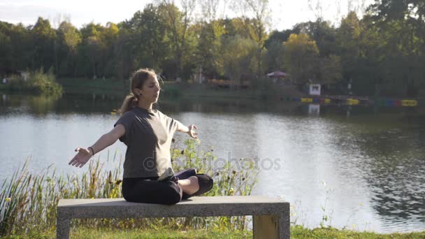 Young adult female practicing yoga in lotus position and having her hands in meditation near to a lake in nature in slow motion — Stock Video