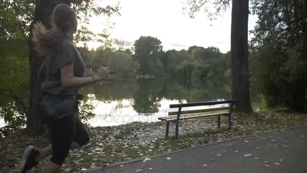 Όμορφη αθλήτρια τζόκινγκ στο πάρκο και να θαυμάσετε τη θέα στη λίμνη κατά τη διάρκεια της ημέρας σε αργή κίνηση — Αρχείο Βίντεο