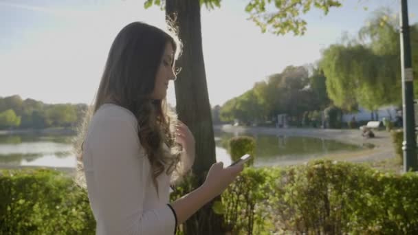 Vacker ung flicka klädd i vit Finkläder för promenader och textning från smartphone i parken i slow motion — Stockvideo