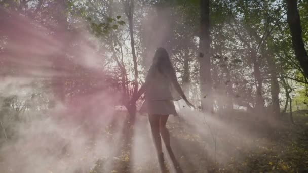 Hermosa joven modelo con un vestido blanco girando en un bosque con humo en un día de otoño en cámara lenta — Vídeo de stock