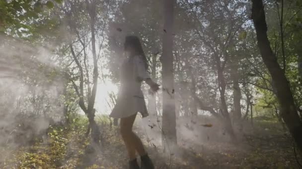 Adolescente chica usando ropa elegante y girando en la luz del sol brillante en un bosque con hojas que caen y humo en cámara lenta — Vídeo de stock