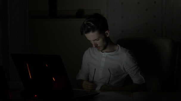 Jeune étudiant confus essuyant ses lunettes tout en utilisant son ordinateur portable à la maison tard dans la nuit 