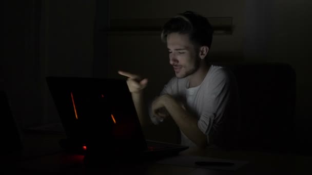 Joven estudiante atractivo de vídeo masculino chateando y saludando a la cámara web en una habitación oscura en casa — Vídeo de stock