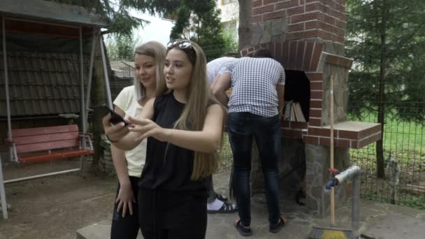 Millennials chicas tomando una selfie con su teléfono inteligente en el patio trasero mientras sus amigos hombres están preparando parrilla de fuego para barbacoa afuera — Vídeo de stock