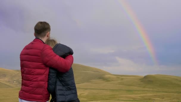 Обнятые мальчик и девочка пара влюбленных любуясь красивым пейзажем с холмами и радугой — стоковое видео