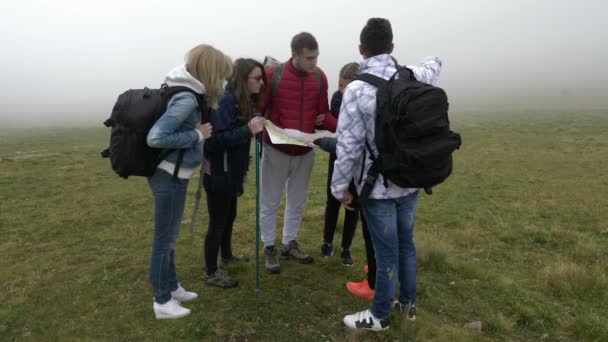 Group of lost young tourists hiking looking for direction trough fog reading the map on a mountain top — Wideo stockowe