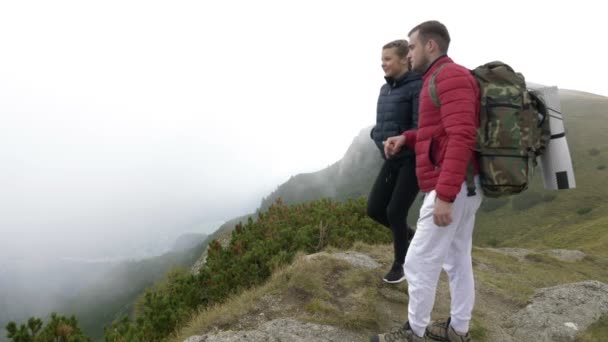 Молодая пара туристов целуются на краю горы наслаждаясь удивительной панорамой долины с лесом, окутанным густым туманом — стоковое видео