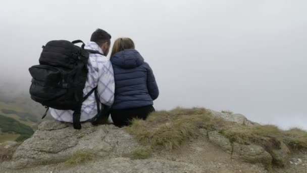 Mladý pár, kteří požívají horské exkurze seděl na vysoké skále a obdivovat krásy přírody s mlžným lesy a zelenými kopci — Stock video