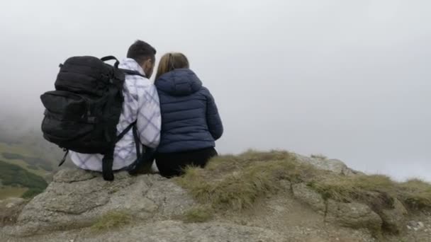 Young couple enjoying the mountain excursion sitting on high cliff and admiring the beauty of nature with misty forests and green hills — Stock Video