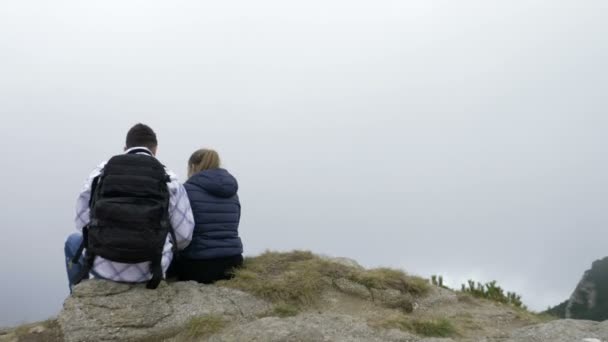 Pareja adolescente sentada en la cima de una montaña rocosa disfrutando de la vista y besándose — Vídeos de Stock