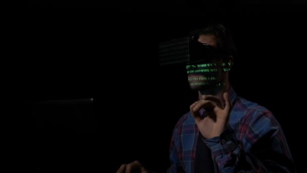 Junger männlicher Hacker mit VR-Brille knackt Codes mit dem Computer aus seinem dunklen Raum, während die grünen Daten auf seinem Gesicht reflektiert werden — Stockvideo