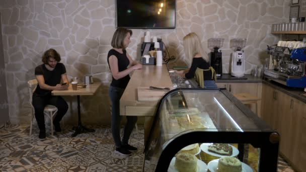 Fröhliche junge blonde Barista-Arbeiterin serviert Kaffee, um zu einer Kundin in einem Café zu gehen — Stockvideo