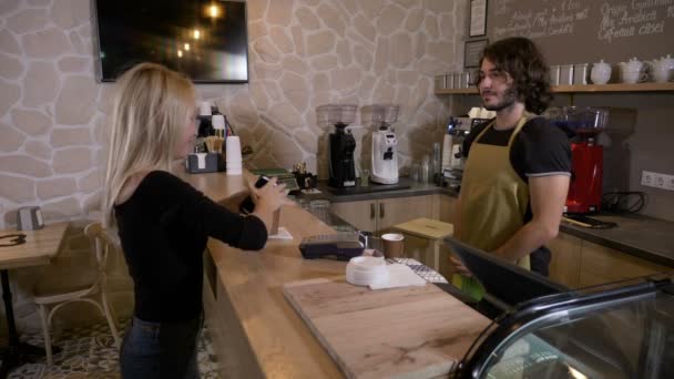 Веселый работник, обслуживающий женщин-клиентов, которые используют приложение для смартфонов и криптовалюту биткойна для оплаты в кофейне — стоковое видео