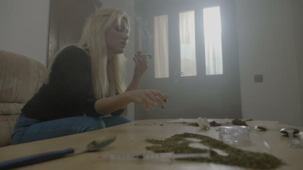 Bored attractive blonde female student sitting on the couch with table full of weed rolling herself a joint lighting and smoking it — Stock Video