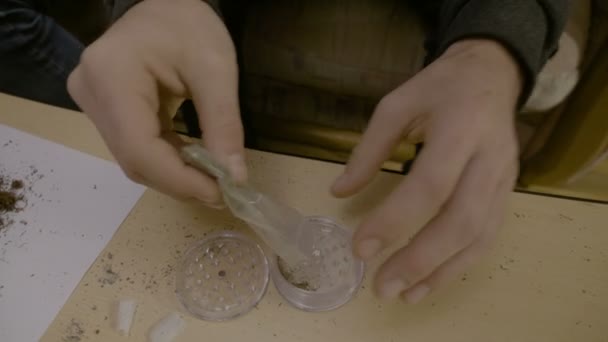 Close up of a man sitting down and grinding marijuana on the table using grinder then rolling in the paper to make a thin weed joint — Stock Video