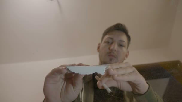 Vista desde abajo de un joven cortando y inhalando cocaína en polvo en casa sobre una mesa de vidrio transparente — Vídeo de stock