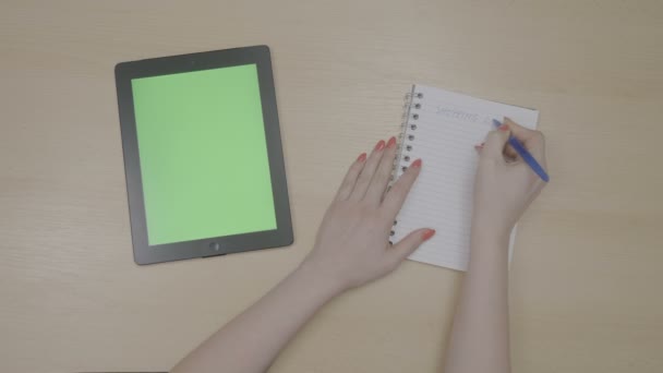 Vista superior de las manos de la mujer planeando lista de compras para las vacaciones con la tableta de pantalla verde PC y la escritura en el bloc de notas — Vídeos de Stock