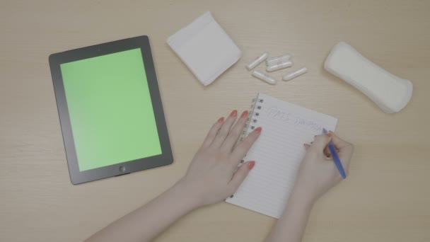 Vista superior de las manos de la mujer con las uñas rojas que planean el síndrome premenstrual Lista del PMS con la PC de la tableta de la pantalla verde y la escritura en el bloc de notas mientras que tiene tampones en la tabla — Vídeos de Stock