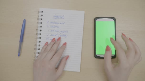 Vista superior de manos femeninas deslizando el teléfono inteligente con pantalla verde mientras mira es la lista de verificación de recetas con ingredientes en su agenda — Vídeo de stock